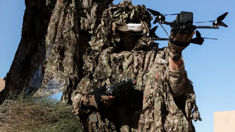 Two soldiers wearing camouflage suits hold a drone and a large gun.