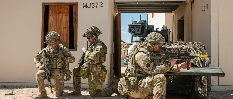 Two British soldiers and an Australian soldier kneel in front of a small green vehicle.