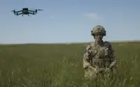 A soldier in camouflage gear stands in a grassy field, holding a controller. A green drone hovers in the sky nearby. The landscape is open with a clear blue sky.