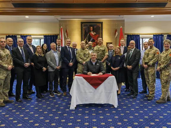 Group photo of military personnel and civilians standing behind a seated man signing a document in a formal room.