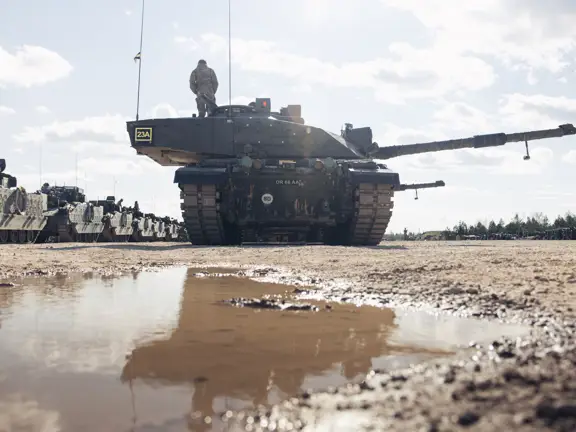 Military tank parked on a muddy ground with a soldier standing on top under a partly cloudy sky.
