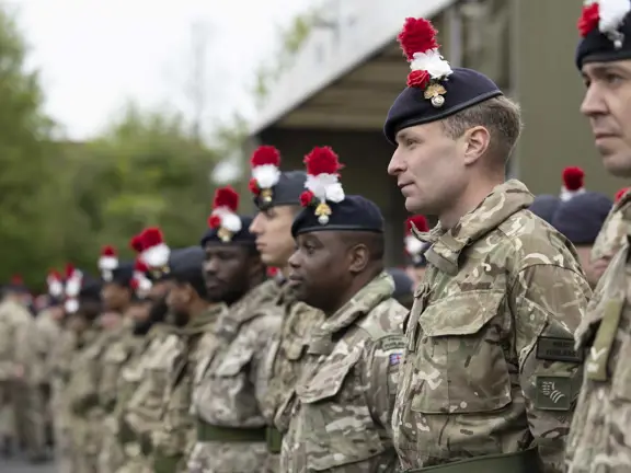 Soldiers in camouflage uniforms stand in formation wearing dark berets with red and white plumes outdoors.