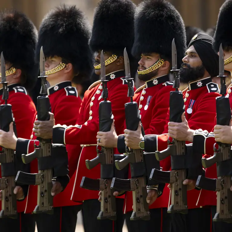 British guards in traditional red uniforms and bearskin hats standing in formation holding rifles.