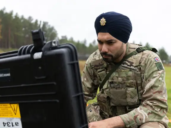 Soldier in camouflage uniform operating equipment outdoors in a grassy field with trees in the background.