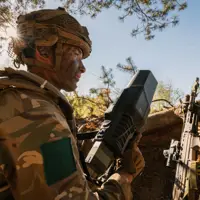 Soldier in camo gear, holding a large device, crouches in a forest setting near a rifle. Sunlight filters through branches, conveying tension and focus.